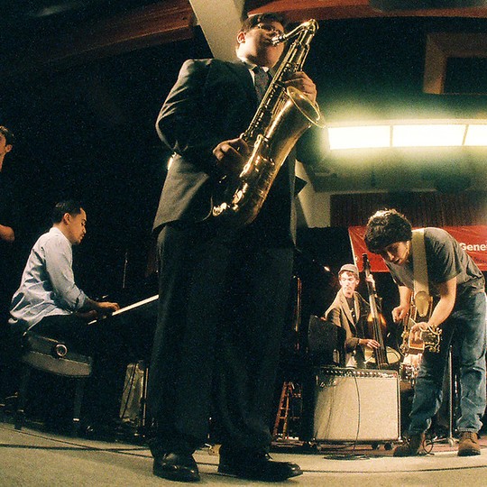 Modern UK jazz musicians playing saxophone piono and guitars live in a smoky scene